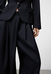 Persona che indossa dei pantaloni larghi neri e un blazer abbinato con un bottone, che mostra una piccola sezione di addome.