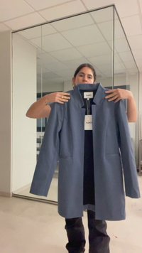 Γυναίκα κρατά ένα μπλε ραμμένο παλτό με λευκή ετικέτα "ONLY" σε ένα δωμάτιο με καθρέπτες σε τοίχους και ταβάνι με πλακάκια.