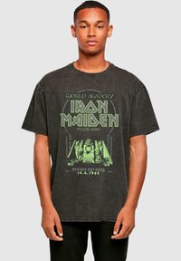 Svart överdimensionerad t-shirt i bomull, med ett grönt grafiskt tryck som inkluderar text och en illustration relaterad till Iron Maidens turné 1985.