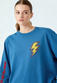 Jeune femme aux cheveux attachés, portant un sweat-shirt bleu avec un écusson jaune en forme d'éclair et un texte rouge sur la manche.
