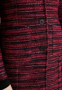 Cappotto intrecciato in rosa e nero con texture, caratterizzato da strisce orizzontali, dotato di bottoni neri e una tasca laterale, che mostra un aspetto ricco e stratificato.
