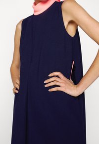 Robe sans manches bleu marine portée sur un col roulé rose, mains sur les hanches, présentant un design ample et minimaliste.