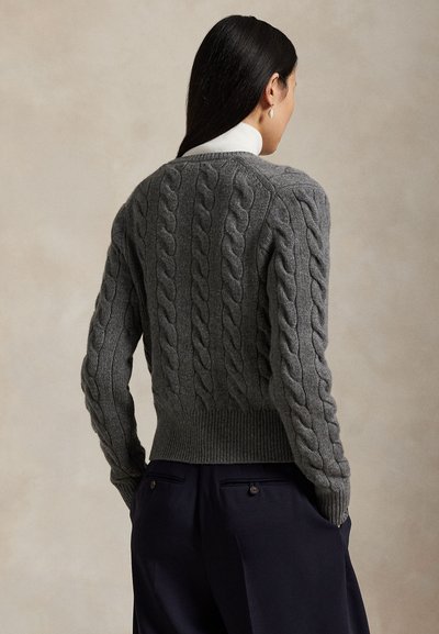 Nő, aki szürke, testre simuló, fonott mintás pulóvert és sötét nadrágot visel, hátulról látható semleges, egyszínű háttér előtt.