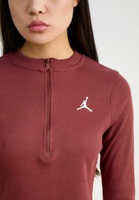 Top lungo a maniche ribattute color marrone bordeaux con colletto con zip, caratterizzato da un logo Jordan bianco sul petto. Tessuto morbido, design aderente.