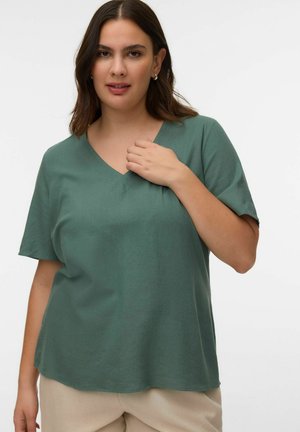 Femeie cu păr castaniu până la umeri, purtând bluză verde cu decolteu în V și mâneci scurte și pantaloni bej, pe un fundal simplu.