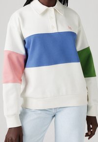 Felpa a polo a maniche lunghe bianca con blocchi di colore orizzontali: azzurro, rosa, verde. Dettagli notevoli includono un colletto e una chiusura con bottoni.