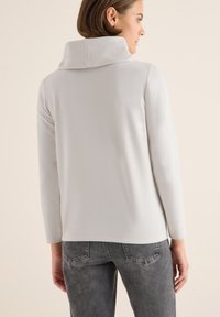 Weißes, langärmeliges Oberteil mit hohem, gefaltetem Kragen. Weicher Stoff und bequeme Passform. Getragen mit grauen Jeans, die ein dezentes Muster aufweisen.