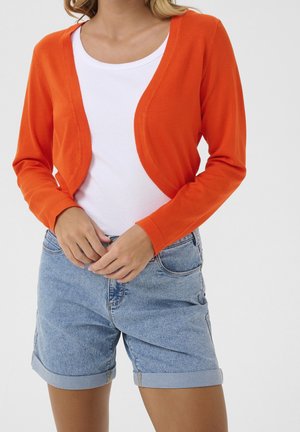 Femme portant un cardigan court orange vif sur un haut blanc, associé à un short en jean bleu clair retroussé.
