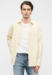 Κίτρινο πουκάμισο με ριγέ σχέδιο και κουμπιά, κατασκευασμένο από ελαφρύ ύφασμα, φορεμένο πάνω από ένα λευκό T-shirt, συνδυασμένο με μπλε τζιν τζιν. Περιλαμβάνει τσέπη στο στήθος.