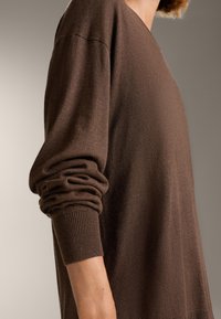 Barna kötött pulóver V-kivágással, laza szabással és bordás mandzsettákkal. Puha textúra és enyhe fényesség látható az anyagon.