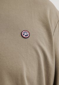 T-shirt di cotone verde oliva con un'etichetta rotonda sul petto, caratterizzata da accenti rossi, bianchi e blu. Tessuto morbido, maniche corte.