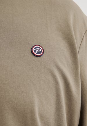 Camiseta de algodón verde oliva con un parche de logo redondo que presenta detalles en rojo, blanco y azul en el pecho. Textura suave, mangas cortas.