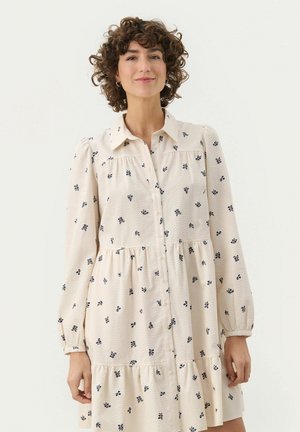 Rochie de culoare crem, cu un material texturat și un model floral în nuanțe de bleumarin, cu guler, închidere cu nasturi pe față și volane pliate. Maneci lungi.
