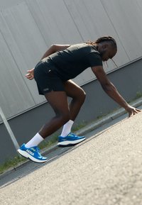 Blauwe hardloopschoenen met witte zolen, bovenwerk van mesh en HOKA-logo; een persoon stretcht op een verharde ondergrond, gekleed in zwarte shorts en witte sokken.