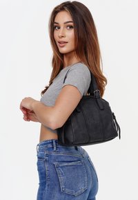 Zwarte leren handtas met een gestructureerd patroon en een ritssluiting. De tas heeft korte handvatten en een gestructureerde vorm, te dragen onder de arm.