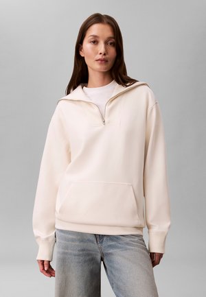 Femme portant un pull crème avec demi-zip et poche avant, associé à un jean bleu délavé, debout devant un fond gris uni.