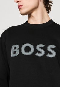 Jeune personne portant un sweat-shirt noir avec un grand lettrage gris « BOSS » sur la poitrine, tête tournée vers la gauche.