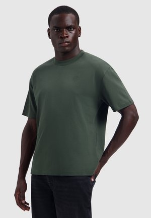 Man wearing a dark green short-sleeve T-shirt with a small tree logo on the chest, standing with one hand in his pocket.