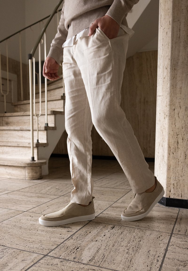 Beige Leinenhosen mit taillierter Passform, kombiniert mit hellen Slippern mit strukturiertem Obermaterial und einer weißen Gummisohle.