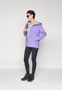 Wodoodporna fioletowa kurtka z szarym wnętrzem, regulowanym kapturem, przednimi kieszeniami, połączona z czarnymi legginsami i butami turystycznymi.