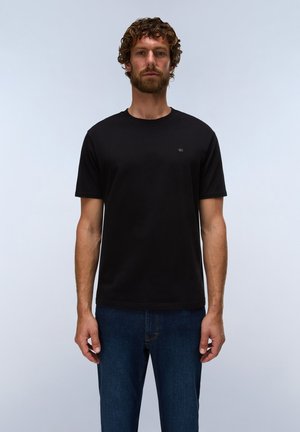 Uomo con capelli ricci e barba che indossa una semplice t-shirt nera a maniche corte e jeans blu su uno sfondo grigio neutro.