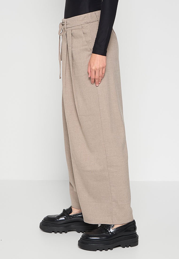 Person, die beige weit geschnittene Hose mit Gürteltrage und schwarze klobige Loafers trägt, steht vor einem schlichten weißen Hintergrund.