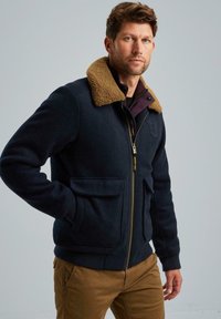 Námořnická modrá bunda s krémovým umělým kožíškem kolem límce, zipovým zapínáním, dvěma předními kapsami, ribbed manžetami a texturovanou tkaninou.