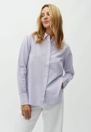 Frau mit schulterlangem blondem Haar, die ein hellviolettes Hemd mit Knöpfen und eine weiße Hose trägt, steht mit einer Hand in der Tasche.