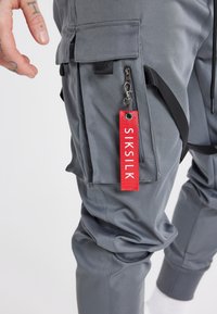 Tummanharmaat cargo-housut, joissa on useita taskuja, vetoketjuja ja punainen SIKSILK-brändäystunniste. Kangas on sileä ja kestävä.