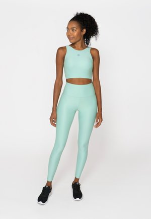 Woman with curly hair wearing mint green sleeveless crop top and high-waisted leggings, standing and looking to her left with black sneakers.