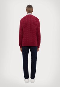 Röd kabelstickad tröja med ribbad mudd och nederkant, bärs med mörka byxor och vita sneakers. Vy från baksidan mot en ljus bakgrund.