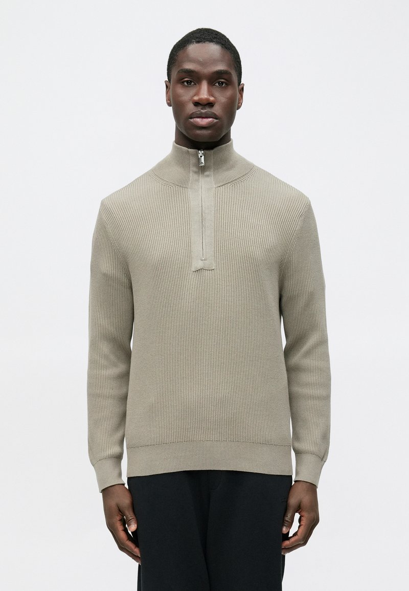 Pull en maille côtelée gris clair avec un col montant à zip et des manches longues, présentant une finition texturée et des poignets ajustés.