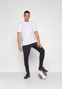 Hvit Nike treningstrøye, svarte slim-fit bukser og svarte fotballsko; står med én fot på en mønstrete fotball.