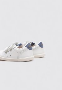 Zapatillas blancas con una parte superior de cuero liso, con un acento azul en el talón, una correa de Velcro y una suela de goma texturizada.