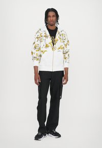White zip-up hoodie with yellow floral pattern, paired with black cargo pants. Black t-shirt underneath, and black athletic shoes.