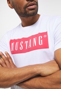 Biała bawełniana koszulka z dużym czerwonym prostokątnym logo z napisem "MUSTANG" w białych, pogrubionych literach na środku.