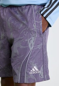 Violetti Adidas-lyhyt housut, joissa on abstrakteja harmaita kuvioita, valkoiset reunat ja käsi taskussa, käytettynä vaaleansinisen hihan kanssa.