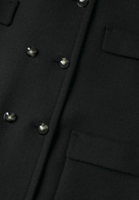 Gros plan sur un tissu noir texturé avec six boutons noirs brillants et deux poches à rabat sur un vêtement croisé.