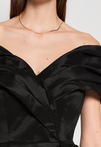 Femme portant une robe noire épaules dénudées avec une texture froncée et un fin collier en or, montrant le décolleté et le haut de la poitrine.