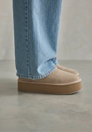 Chaussons en suède beige à plateforme avec une finition lisse, associés à un jean en denim bleu clair. Les chaussures présentent une épaisse semelle marron et un bout arrondi.