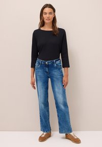 Femme debout contre un mur beige portant un haut noir à manches longues, un jean bleu droit et des baskets marron avec des lacets blancs.