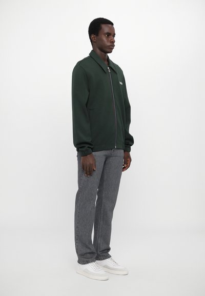 Grønn zip-up hettegenser med krage, grå teksturerte bukser og hvite joggesko. Stitched detaljer på jakken og livet.