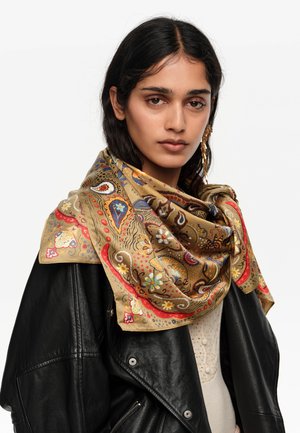 Kobieta o długich ciemnych włosach nosi duży jedwabny szal w złote, czerwone i niebieskie wzory, zarzucony na czarną skórzaną kurtkę i beżową bluzkę.