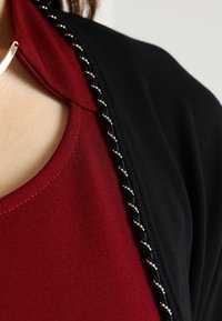 Chal negro con acentos de cuentas plateadas, superpuesto a una blusa burdeos. La tela parece suave con un diseño drapeado.