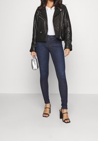 Chaqueta de motociclista recortada de cuero negro, camiseta blanca, jeans ajustados azul oscuro y sandalias de tacón alto negras con tiras. Bolso plateado en la mano.