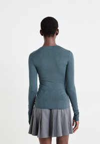 Haut à manches longues teal avec une coupe ajustée, associé à une jupe grise plissée. Texture lisse ; aucun motif ou équipement visible.