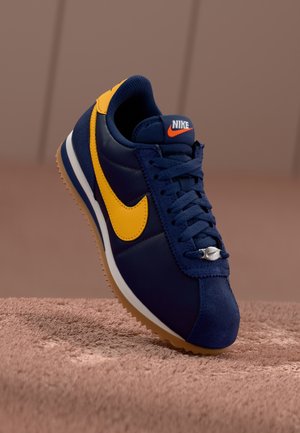 Marineblå og gul Nike sneaker med gummisål, detaljerede snørebånd og Nike-logo på tungen og siden, vist mod en brun baggrund.