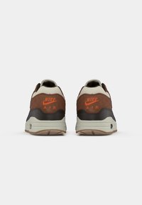 Um par de ténis Nike Air castanhos e bege vistos de trás, com o logótipo da Nike em laranja e uma entresola branca, sobre um fundo branco.