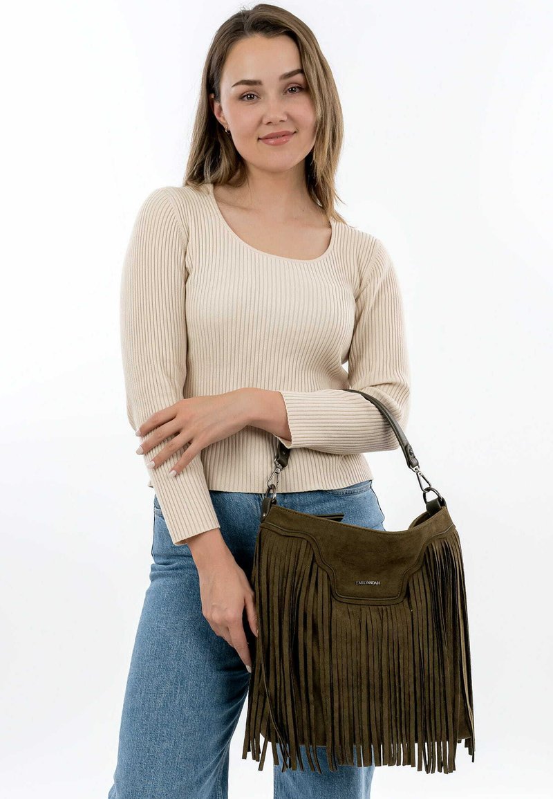Dark green suede handbag with fringe detailing, short strap, and a structured body. Model wears a cream ribbed top and light blue jeans.