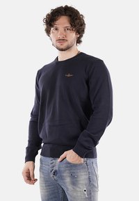 Maglione lavorato a maglia blu navy con collo rotondo, polsini e orlo a costine, caratterizzato da un piccolo emblema dorato sul petto.